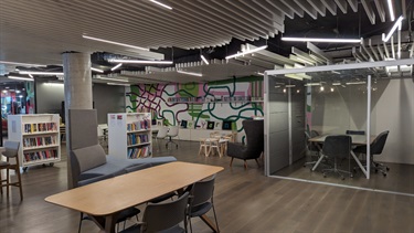 Photo shows the YARN interior, positioned in the right hand corner furthest from the entrance. There is the wooden table in the foreground of the frame and the T shaped sofa to the left of the table. The enclosed working space is situated on the right hand side of the frame and it has glass sliding doors and a table with 6 chairs around it. In the background you can see the childrens tables and the backs of the armchairs, as well as the wooden benches that skirt the border of the left hand side of the room (as you enter), there are cushions along the benches that spell out THE YARN with each letter printed on a separate pillow.