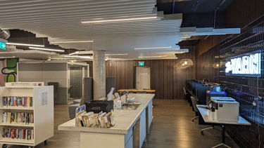 Photo shows the YARN interior, positioned in the right hand corner closest to the front entrance. In the foreground of the middle of the frame is the kitchen bench space. situated behind the kitchen bench is the T shaped sofa and to the left of that is are the bookshelves. To the right of the kitchen bench is a table that holds the paper printer and the 3D printer. The computer cubicle stations are running along the right hand side wall. The emergency fire exit door is pictured on the back wall (further from the front entrance).