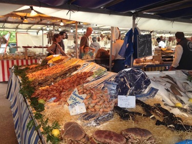 Photo shows the Le Vesinet fish markets. The closest stall has a wide range of seafood types, incoluding shrimp and crabs, as well as fish. You can see a family browsing the goods and chatting to the proprietor.