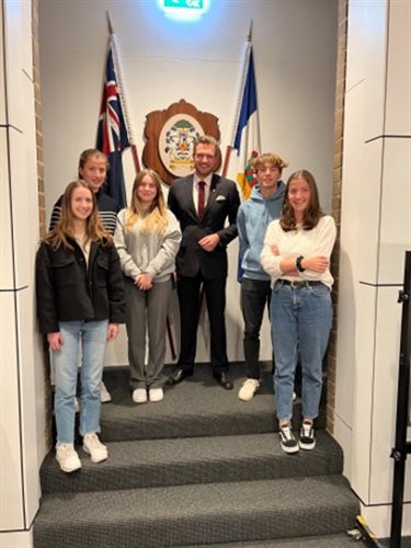 Photo shows the Le Vesinet exchange students standing with Mayor Miles in the Council chambers. There are 5 students and they are all standing together underneath the Australian flag and the Hunters Hill Coat of Arms.