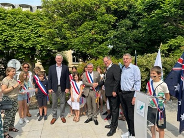 Photo shows Councillors Ross Williams and Richard Quinn visiting le Vesinet. They are being greeted by community representatives and children. They are standing together in a semi-circle. behind them are some very lush trees and there is a yellow building barely visible through the trees.