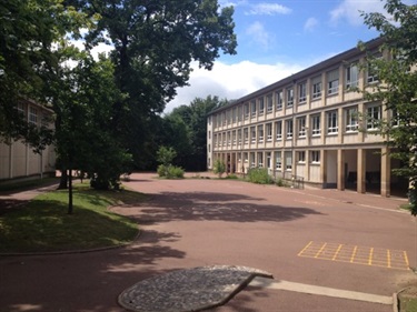 Photo shows the grounds of the high school that is home to the french students and temporary home of the australian students. it shows a large paved courtyard. To the left of the courtyard are a series of trees that shade the area. To the right of the courtyard is a large rectangular building with many windows.