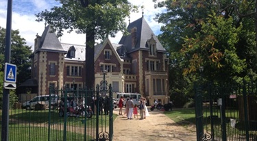 Photo shows a large Château with a number of people walking towards it. There are cars in the forecourt area and there are metal gates in front of the property grounds. Surrounding the property are a number of very tall trees. The building is made of brick/stone and the roof is heavily slanted. There are many windows surrounding the entire exterior of the property, at least two stories tall.
