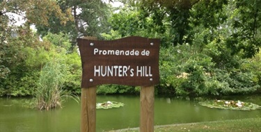 Photo shows a sign situated in front of a small river. Sign says Promenande de Hunters Hill. behind the river is lots of vegetation and there are lovely floating lilly pads on the water.