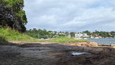 Photo is of the waters edge as accessed via the bushwalk. From the middle of the frame, at the horizon line and extending to the right of the frame, is the Parramatta river, with a number of sailing boats and apartment dwelling situated behind it. The foreground is of sandstone rocks with some small puddles in the rock indents. Surrounding the rocks is grass and there is a tree visible in the left of the frame.