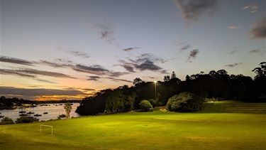 Bedlam Bay oval illuminated by floodlights, facing towards the water with the setting sun in the background