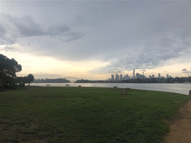 Photo shows a wide angle of the tip of the reserve, showing the spectacular panoramic view of Sydney Harbour. the harbour bride sits far in the background and the sky is overcast with sunlight pouring through.