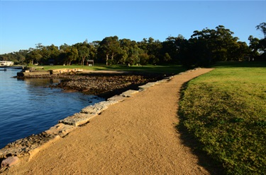 Gravel footpath flanked by grass and travelling along the harbour foreshore. Abundant trees in the background