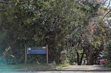 Photo is of the entrance pathway and sign as seen from the street. The sign is positioned to the left of the access pathway, which is on the right side of the frame. Behind the entrance sign is dense vegetation that is very tall (several metres) and occupies the vast majority of the shot. The road is just barely within frame and there is some sunlight distortion in the bottom left corner..