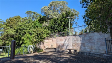 Photo shows the Joubert St Sundial. The sundial is situated to the left of the frame, as is well shaded by nearby trees (situated behind the camera operator). To the centre/right of frame is a small sandstone retaining wall that guards the steps that lead down from Joubert Street. The sky is entirely clear and bright. There is heavy tree coverage situated in the background, set down from the sundial platform.