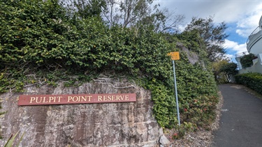 Photo shows the entrance sign that is attached to the rockface surrounding Pulpit Point Reserve. On the right hand side of the frame you can see the pathway that leads around the rockface and grants entry to the reserve.
