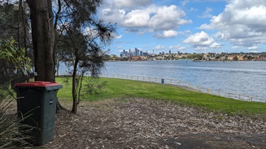 Photo shows the view that greets you as you enter Pulpit Point Reserve. Shows the public red bin on the left of the frame. To the right of the frame is the Parramatta River and in the background is a view of Sydney City as well as Spectacle and Cockatoo Islands.