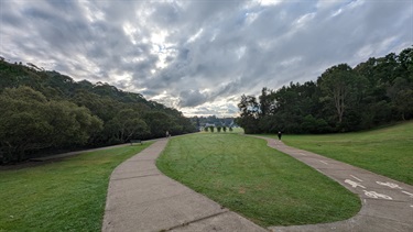 Photo shows a very wide angle view of the cyclist and foot traffic pathway. The pathway forks into a left and right option, both eventually leading back around to the start. Extensive storm clouds can be seen in the sky but the sunlight is illuminating the outer edges. The left pathway leads down next to the water whereas the right pathway leads down past more trees.