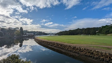 Photo is taken from the Joly Parade Footbridge, shows the Tarban Creek on the left hand side and a view of the Riverglade Reserve soccer pitches on the right hand side. Gladesville Bridge visible in the background and an extensive sky with clouds visible in the top of frame.