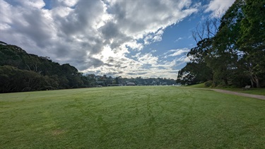 The first of two soccer pitches, wide angle shot showing the wide open space, flanked by trees on the left and right, showing dwellings very far in the background, as well as a sky with overcast clouds, but still lots of blue.