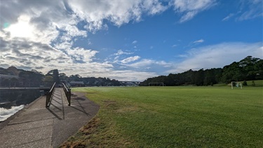 A wide angle photo showing the second (larger) soccer pitch, as well as the Joly Parade Footbridge (which allows access to/from the reserve). The sky is exceptionally large and features overcast clouds on the left hand side and beautiful clear blue sky on the right hand side. The Gladesville Bridge is just barely visible in the background.