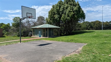 Photo shows the practice basketball hoop that sits next to the toilet block. There is lush green grass surrounding both structures and there is desh vegetation positioned behind them.
