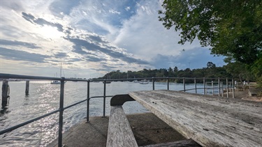 A bench sits in the bottom right corner, extentin to the middle of the frame. The water sits beyond the hand railing, which surrounds the stone platform. The sky occupies almost the entire upper half of the frame, slightly overcast with strong sunlight shining through, the far right-upper half of the frame contains a tree canopy. The background shows boats floating on the water, as well as mangrove and bush lands.