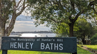 photo shows the Henley Baths entrance sign. The sun is shining strongly off the water, which sits in the background. The Sign sits promonently in the foreground and there is lush grass all around. There are two trees within the reserve and several in a nearby private garden to the right. There are buildings in the far background beyond the water.