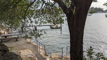 Photo has a warm glow to it. There is a tree in the foreground, offset to the right. Low in the frame is the stone platform. Offset to the left, on the stone platform, there is a wooden table with benches for 6 people. The water is gently lapping at the cut stones that serve as the entry to the water. There is a private jetty in the background and the far background is mostly obscurred by the aformentioned tree. The right of frame is the wider river.