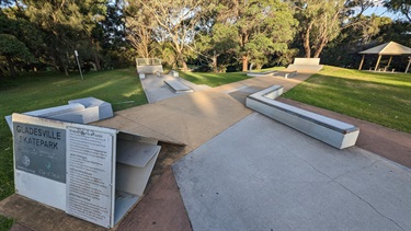 Photo shows the Gladesville Reserve Skatepark from a high angle, positioned with Victoria Road to the back of the photographer. There is a 35 degree ramp on the lower-left side of the frame. There is are four concrete platforms that are around .5 metres tall each, they are arranged evenly around the skatepark. There is a steep ramp positioned far away, in the middle of frame, with a grinding rail leading up to the ramp. To the far right of the frame is an additional ramp, with a sheltered gazebo set to the right of said ramp.