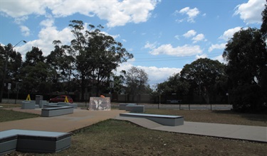 Photo shows the Gladesville Reserve Skatepark from eye level, facing towards Victoria Road. There are numerous concrete platforms distributed evenly around the skatepark, as well as paved areas between all sections. There is the inclined ramp set amongst the platforms. Behind the park is Victoria Rd and the nearby Huntleys Cove development area, including many large trees.