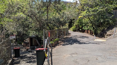 Photo shows a path that forks in two directions. To the left leads down towards the water and the right fork leads up to Collingwood Street. At the fork are two red general waste bins for the bath goers to use.