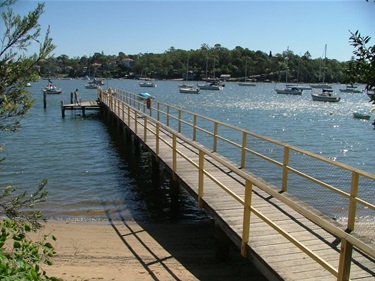 Photo shows the netted area of the Woolwich Baths. Shows a walkway that juts out from the shore and has a small standing platform at the end. The sun is clittering off the water. You can see a swimming standing at the end of the walkway.