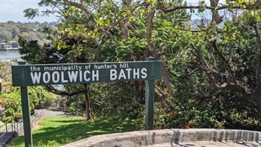 Photo of the woolwich baths entrance sign. To the left of the sign are the steps that lead down to the baths area. To the right is some dense vegetation and behind the sign is beautiful lush grass. Left side background shows the Lane Cove River.