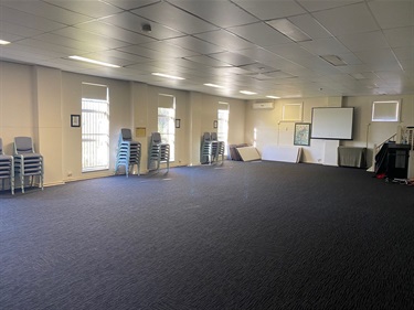 Photo shows the interior of Fairland Hall. The floor is is carpet. On the left hand of frame is a wall that has a set of three windows (more windows are out of frame). There are plastic chairs stacked in small piles along the wall with the window. On the right hand side of the frame you can see a projector screen and some tables stacked against the wall. The right hand side wall also has small windows that have the blinds drawn down.