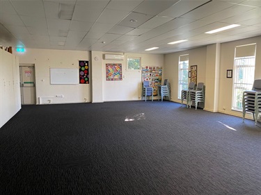 Photo shows the interior of Fairland Hall. The right hand side of the frame shows two large windows in the wall, as well as stacked chairs along the same wall. There is some colourful community group quilts hung up on the wall on the right hand side of the frame. There is a whiteboard situated along the far wall, slightly to the left of frame. The fire escape door is situated to the left of the whiteboard. The far left of frame shows some of the wall cupboards.