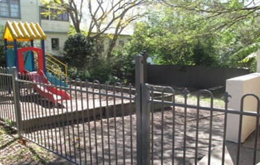 Photo shows the small playground that is situated to the left of the front entrance. The area is fenced off and the play equipment consists of a small climbing frame with a slide and a small staircase. The playground is sitting atop of a raised platform. The background of the photo shows a double storey dwelling that neighbours Fairland Hall.
