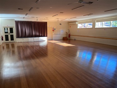 Photo shows the main floor area, facing towards the stage. The floor is made of wood. There is a fire extinguisher in the opposite corner to where the photographer is standing. There are small square windows that are in sets of three, two sets are present on the right hand side of the picture. There is an air conditioning unit situated to the left of the windows. To the left of the air con unit is a doorway that leads to the kitchen area. To the left of the doorway is the stage area, curtains drawn which obscurs the rest of the stage. To the left of the stage is a set of two doors, which lead to the front entrance. To the left of that door is another fire extinguisher.