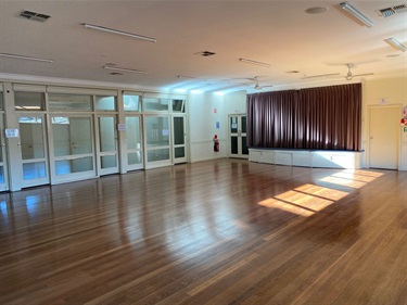 Photo shows the community hall from the other side of the room to interior 1, also facing towards the stage area. Instead of the windows on the far wall, this photo shows the glass door flanked walking corridor that links to the front entrance area.