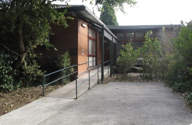 Photo shows the front entrance of Gladesville road community centre. There is a ramp that extends from the bottom left of the photo frame, up towards the centre-right of the picture, where it meets the front door to the community hall. The hall is made of brick and has a corregated metal roof, with overhanging awnings sheltering the entrance area. There are a few windows situated on each wall, as well as a few fluro lights. There is a paved area that can be used to park a mobility assistance vehicle. Trees and bushes surround the paved area and the community hall.