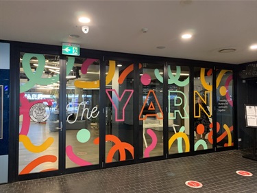 Photo shows the YARN exterior glass wall and front entrance door. The glass has colourful swirl like designs and THE YARN is printed in large lettering across the length of the glass wall. You can see into the interior of the yarn and one of our staff members is in there setting up for the day ahead.
