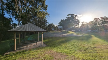 Photo shows the Gladesville Reserve basketball court. left of frame is a gazebo with bench table. Mid-frame is the basketball court. Right of frame is the setting sun and Victoria Road traffic.