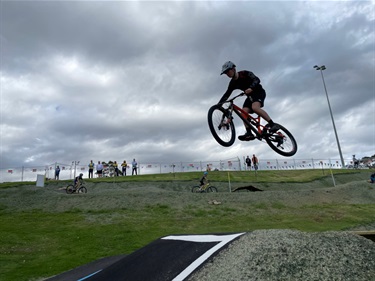 Photo shows a young man taking a very high jump, travelling right to left.. Photo is from a low angle and the rider is set against the overcast sky. There is a hill in the mid/background and it shows a set of eight people watching over the riders. There are also two other riders on the higher section of the track, set directly below the observes at the top of the hill.