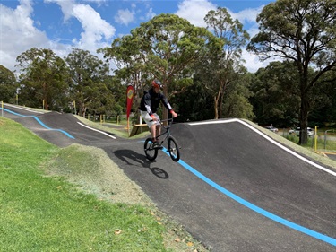 Photo shows a young man coming down from a jump and he is performing a wheelie upon landing, travelling left to right. His face is focused and determined. To the riders left is the section of track that he just came from, it is a series of 