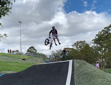 Photo shows a rider performing an excellent trick whilst going over a very high jump. He is travelling right to left in the frame. His hands are firmly gripping the handle-bars and his legs are both extended downwards, he appears to have kicked the bike frame around in a 360 degree rotation. His eyes are focused firmly on the landing zone. Behind the first rider, to the bottom right of frame, is a second rider. The second rider is also performing a jump. To the left of both riders is the hill, upon which a few observers are standing. There is also another rider/adult standing in the bottom right of frame. The sky is set with large overcast clouds. There are trees in the background.
