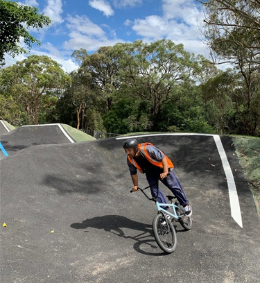 Photo shows a gentlemen taking a tight corner around the track. He is travelling right to left. His eyes are pointing in the direction he is turning and he is very focused. He is standing high on the bike pedals to acquire additional speed. In the background is the extensive Boronia Park bushland and the sky is sunny with mild clouds.