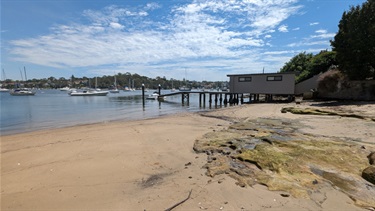 Photo shows the un-netted area of the baths. There are large flat rocks to the right of frame, set amongst sand that sits next to the water on the left of the frame. There are lots of boats out on the water. There is a boat dock set in the background, as well as lush mangrove and bushland on the other shores of the river.