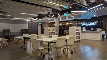 Photo shows the YARN interior, positioned in the left corner closest to the doorway. There are two tables with seating capacity of 6 each. There are very small childrens tables and chairs in the left of frame. Behind all of the tables are two large grey armchairs with high backs. To the right of frame are the two bookshelves and behind them (on the right hand wall) is the kitchen area and the lit up sign for THE YARN.