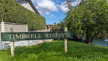 Photo shows the entrance to Timbrell Reserve. The grass is very lush and the sky is mostly clear with only a few clouds. You can see the Parramatta River in the bottom right of the frame.