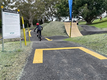 Photo shows a rider who has just set off from the starting line. The rider is travelling away from the camera and his bike is tilted as he is picking up speed. To the riders left is a large sign explaining the rules of the track. To the right of the rider is a very steep wooden jump and to the right of the jump is a couple taking photos. The background is off the bushland adjacent to the track + some of the lush grass that fills out Boronia Park.