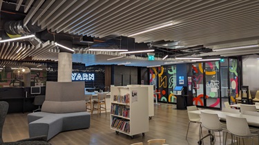 Photo shows the YARN interior, positioned in the left hand corner furthest from the door. It shows the adult sized tables to the right of frame and the bookshelves in the middle of frame. To the left of the frame is the T shaped sofa and behind that is the kitchen bench. You can see the front entrance in the background.