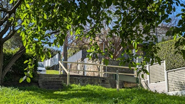 Photo shows Timbrell Reserve from the perspective of the bench looking back into the interior of the reserve. Shows very lush grass, with the small stone wall/steps that sit between the reserve and the street. You can see green leaves coming down from the top of the frame and they are well illuminated by sunlight. You can see the bright sky in between the leaves. There is a nice stone dwelling set on the street, in the background.